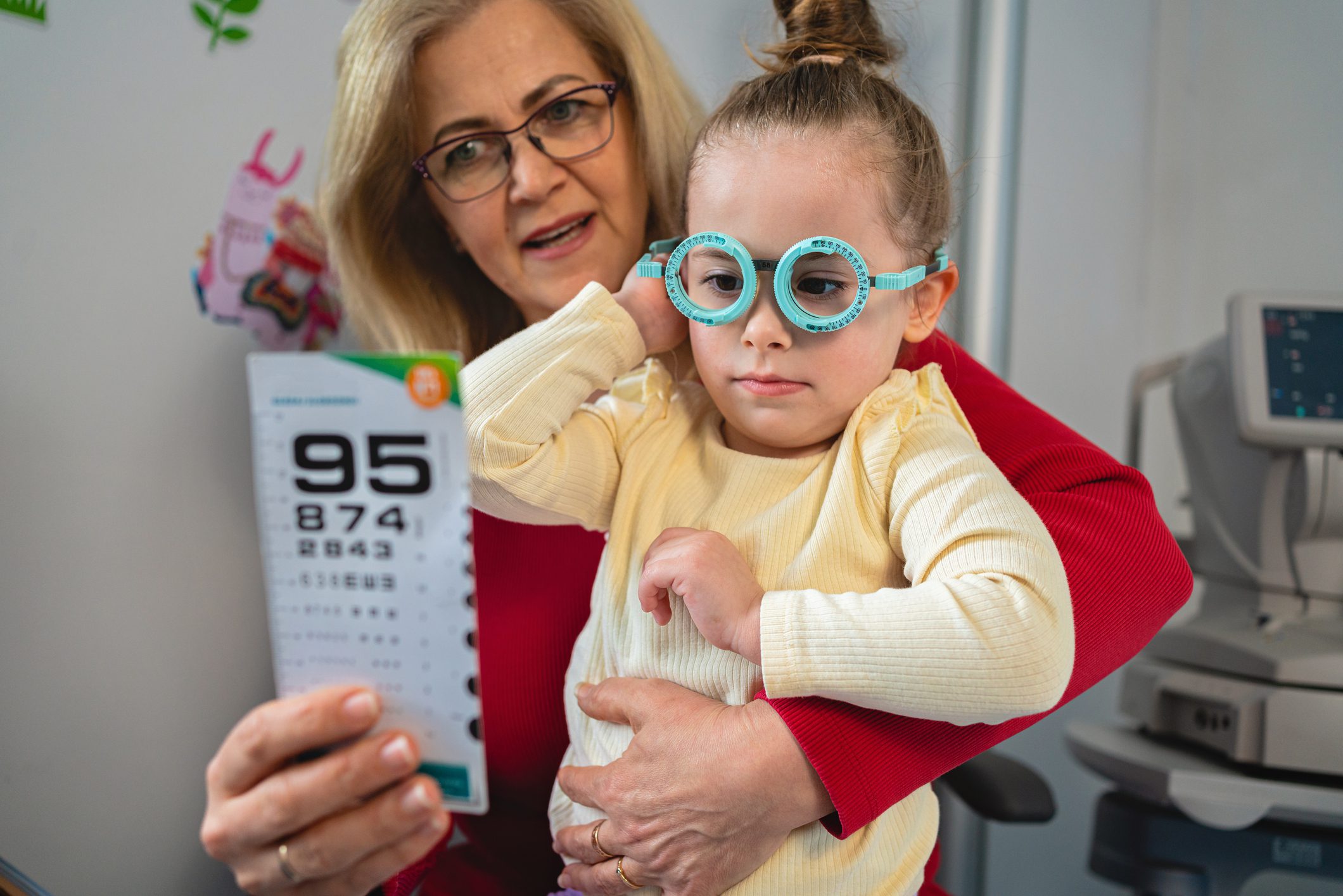 A child with glasses is held by an adult while holding an eye test chart.