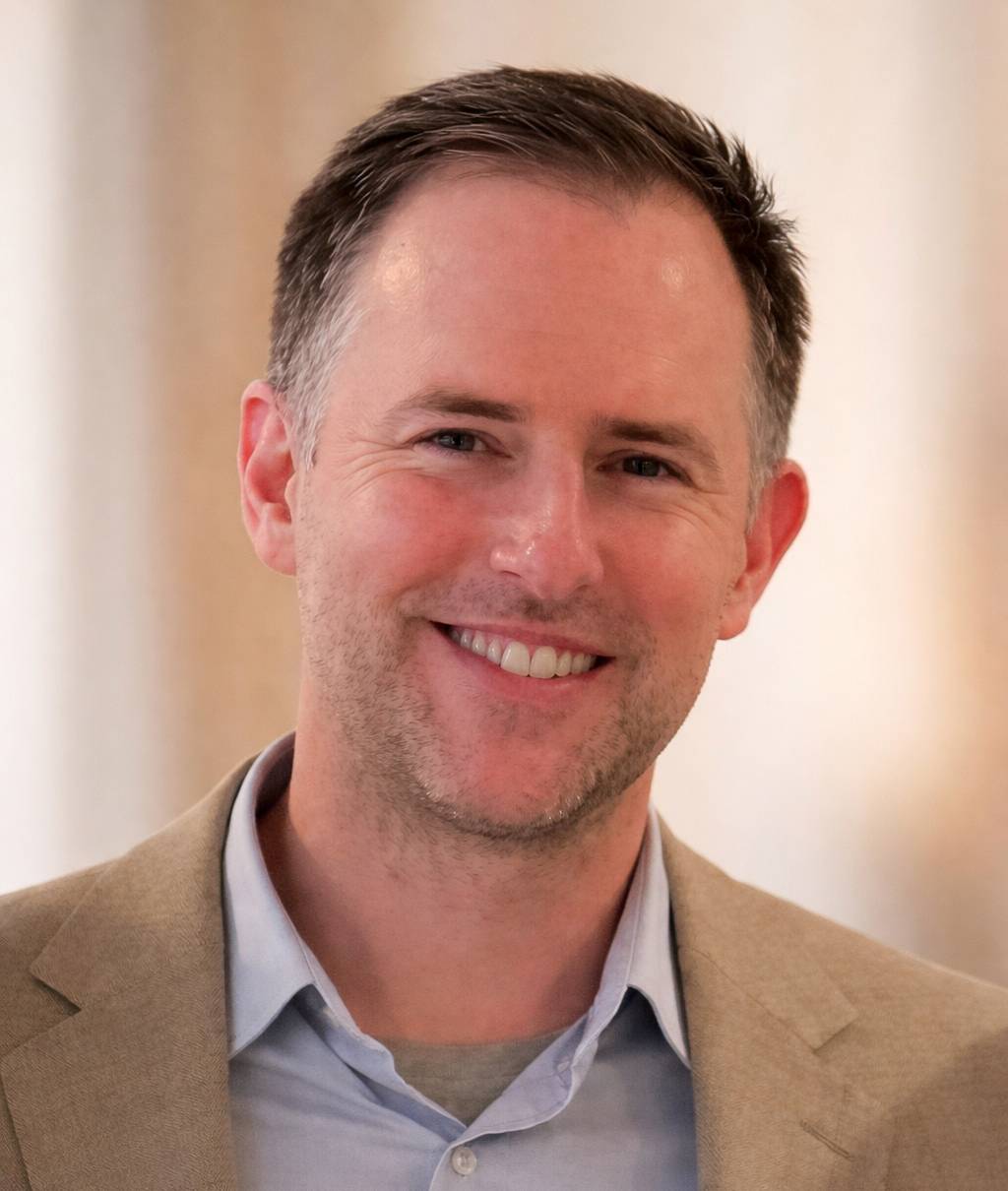 Smiling man in a beige blazer with a light background.